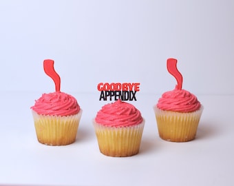 May include: Three pink frosted cupcakes with red and black decorations. The middle cupcake has a topper that says "GOODBYE APPENDIX".