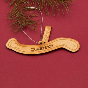 May include: Wooden Christmas ornament shaped like a curved bone with the text "COLLARBONE 2024" and a separate tag with the text "YOUR NAME".