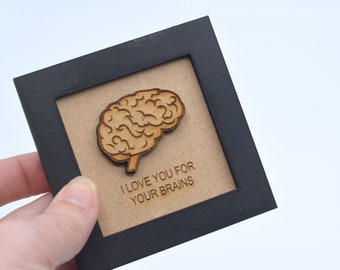 May include: A small framed wooden sign with a brown background. The sign features a wooden cutout of a brain and the text "I love you for your brains".