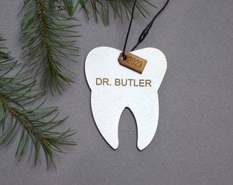 May include: A white tooth-shaped ornament with the text "DR. BUTLER" and a small wooden tag that reads "2023". The ornament hangs from a black string and is set against a gray background with a sprig of evergreen.