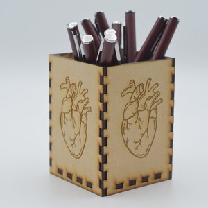 May include: A wooden pencil holder with a heart design. The holder is brown and has a black border. It is filled with red pens.
