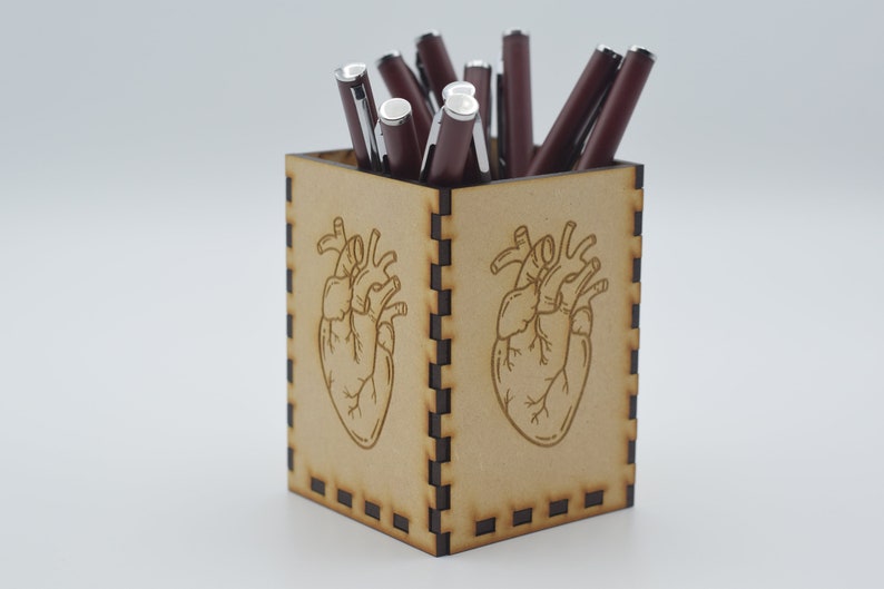 May include: A wooden pencil holder with a heart design. The holder is brown and has a black border. It is filled with red pens.