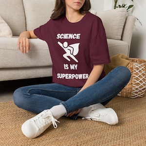 May include: A maroon short-sleeved t-shirt featuring a white graphic of a stick figure wearing a cape in a flying pose, with the text "SCIENCE IS MY SUPERPOWER" printed in white block letters across the chest, paired with blue denim jeans.
