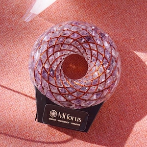 May include: A spherical, amethyst crystal sculpture with copper wire detailing. The sculpture has a central opening and sits on a black base with a label that reads "Mi Torus". The background is a textured, reddish-pink fabric.