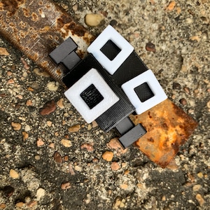 May include: A black and white geometric accessory with a rectangular shape. The accessory features a series of white squares with black square centers, connected by black bands. The object is placed on a rusty metal surface.