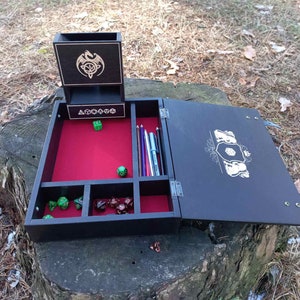 May include: A black wooden box with a red felt interior and compartments for dice and pencils. The box has a hinged lid with a decorative design and a separate compartment for dice.