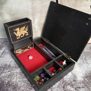 Könnte beinhalten: Eine schwarze Würfelbox aus Holz mit einem Klappdeckel und mehreren Fächern. Die Box hat einen rot mit Filz ausgekleideten Rollbereich und einen separaten Turm. Goldene Drachen- und Schlangenmotive sind auf die Box geätzt. Verschiedene Würfel und Stifte sind im Inneren sichtbar.