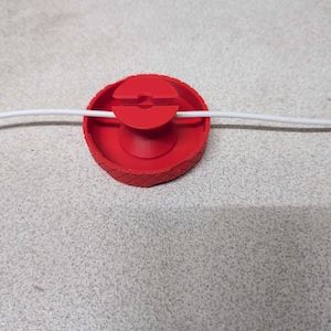 May include: A red 3D-printed cable organizer with a grooved edge and a central slot. A white cable is threaded through the organizer. The organizer is designed to manage and secure cables.