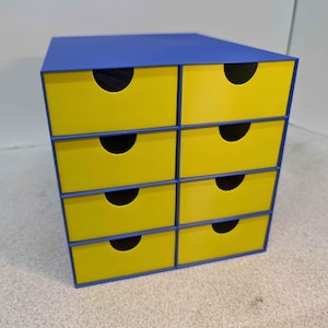 May include: A small, six-drawer storage unit. The top is blue, and the drawers are yellow with black, semi-circular cutouts for handles. The unit is a cube shape and sits on a light-colored surface.