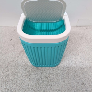 Op de afbeelding: Een turquoise plastic container met een witte rand en deksel. De container heeft verticale geribbelde details aan de buitenkant. Het deksel is open en onthult de binnenkant van de container.