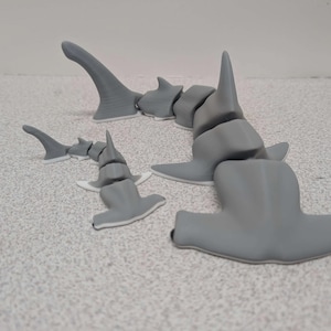 May include: Gray articulated hammerhead shark model with a segmented body and fins. The model is 3D-printed and designed to be poseable. The shark is displayed on a speckled surface.