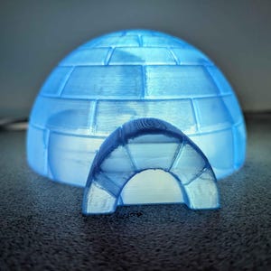 Puede incluir: Un modelo de iglú azul brillante con una entrada a juego. El iglú está hecho de un material translúcido, con patrones de bloques visibles. El interior está iluminado, creando un brillo suave y etéreo. Es un artículo decorativo.