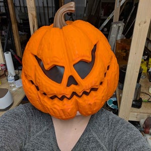 Op de afbeelding: Een oranje pompoenmasker met een uitgesneden gezicht, met grote ooggaten en een grillige mond. Het masker heeft een bruine steel en wordt gedragen door een persoon die een grijze trui draagt. Een Halloween-kostuum.