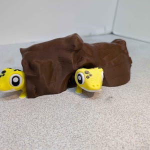 May include: A brown, cave-like structure with two yellow lizard figurines. The lizards have black spots and large, round eyes. The figurines are positioned near the cave, suggesting a playful or decorative theme.