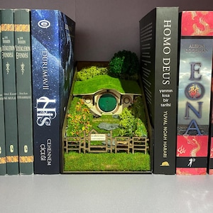 Könnte beinhalten: Ein Miniatur-Hobbit-Loch-Diorama mit grüner Tür und Dach, umgeben von künstlichem Gras und einem Holzzaun. Das Diorama befindet sich zwischen mehreren Büchern auf einem Regal und schafft eine skurrile Präsentation.