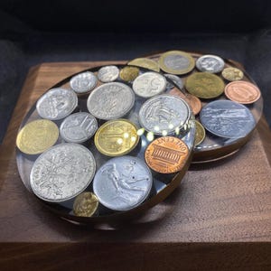 May include: Two round coasters made of clear resin with a variety of coins embedded inside. The coins are mostly silver and copper in colour, and some have a gold hue. The coasters are on a wooden surface.