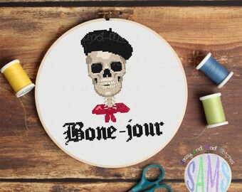 May include: A white cross-stitch embroidery hoop with a skeleton wearing a red bow tie and a black beret. The text "Bone-jour" is stitched below the skeleton.