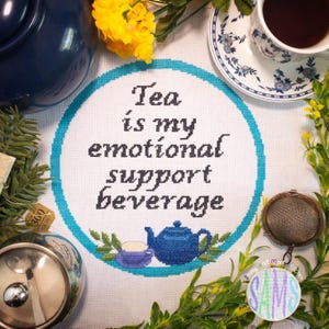 May include: A cross-stitched design with the text "Tea is my emotional support beverage" in black lettering, surrounded by a teal circle. The image includes a blue teapot, teacup, and tea-related accessories, with floral accents.