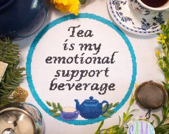 May include: A cross-stitched design with the text "Tea is my emotional support beverage" in black lettering, surrounded by a teal circle. The image includes a blue teapot, teacup, and tea-related accessories, with floral accents.