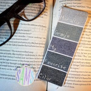Pode incluir: Um marcador de página azul, cinza e branco. O marcador tem as palavras "Minha cor favorita" bordadas. O marcador está em cima de um livro.
