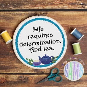 May include: A blue and white embroidery hoop with the text "life requires determination. And tea."  The hoop is surrounded by a wooden background with a pair of scissors and spools of thread.