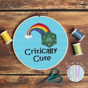 Pode incluir: Um aro de bordado azul com um arco-íris, um dado verde de 20 faces e o texto "Critically Cute".