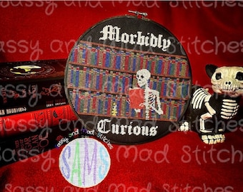 Könnte beinhalten: Ein schwarzer und weißer Kreuzstich-Stickrahmen mit dem Text "Morbidly Curious" und einem Skelett, das ein Buch in einer Bibliothek liest. Der Rahmen befindet sich auf einer roten Oberfläche mit einer schwarzen und weißen Skelettkatzenfigur.