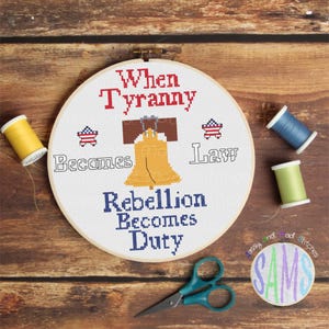 May include: A white embroidery hoop with a cross-stitch design featuring a Liberty Bell, stars, and the text "When Tyranny Becomes Law Rebellion Becomes Duty".