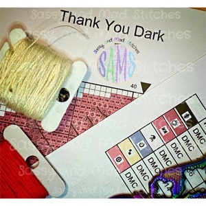 May include: A close-up of a cross-stitch pattern with the text "Thank You Dark" and "Sassy and Mad Stitches". The pattern includes a circle with the letters "SAMS" in different colors. The image also shows a spool of white thread, a spool of red thread, and a needle.