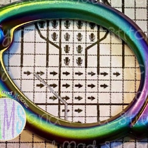 May include: A close-up of a rainbow-colored metal hoop with a star design. The hoop is on a gridded surface with arrows pointing to the right. The text "Cross Stitch" is visible on the surface.