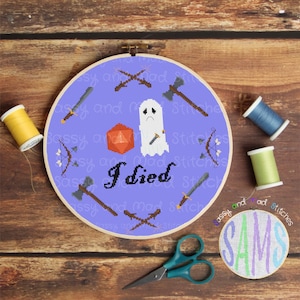 May include: A purple cross-stitch embroidery hoop with a white ghost holding a dagger, a red 20-sided die, and the text "I died". The hoop is surrounded by various weapons, including swords, axes, and bows and arrows.