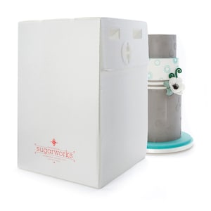 May include: A white corrugated cardboard cake carrier with a locking mechanism. The carrier has the red text "sugarworks" printed on the side. A three-tiered gray cake with teal accents and a white flower is visible in the background.