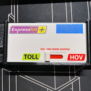 May include: A black plastic device with a white faceplate and a red and white toggle switch. The faceplate has the text "ExpressToll+" in purple and yellow, "TOLL" in green, "HOV" in red, and "HOV - ONLY WHERE ACCEPTED" in black.