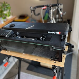 Puede incluir: Un entrenador de bicicleta Sparx negro con una cubierta de plástico transparente, montado en un banco de trabajo de madera con abrazaderas. El entrenador está diseñado para sujetar una bicicleta en su lugar para el ciclismo en interiores.