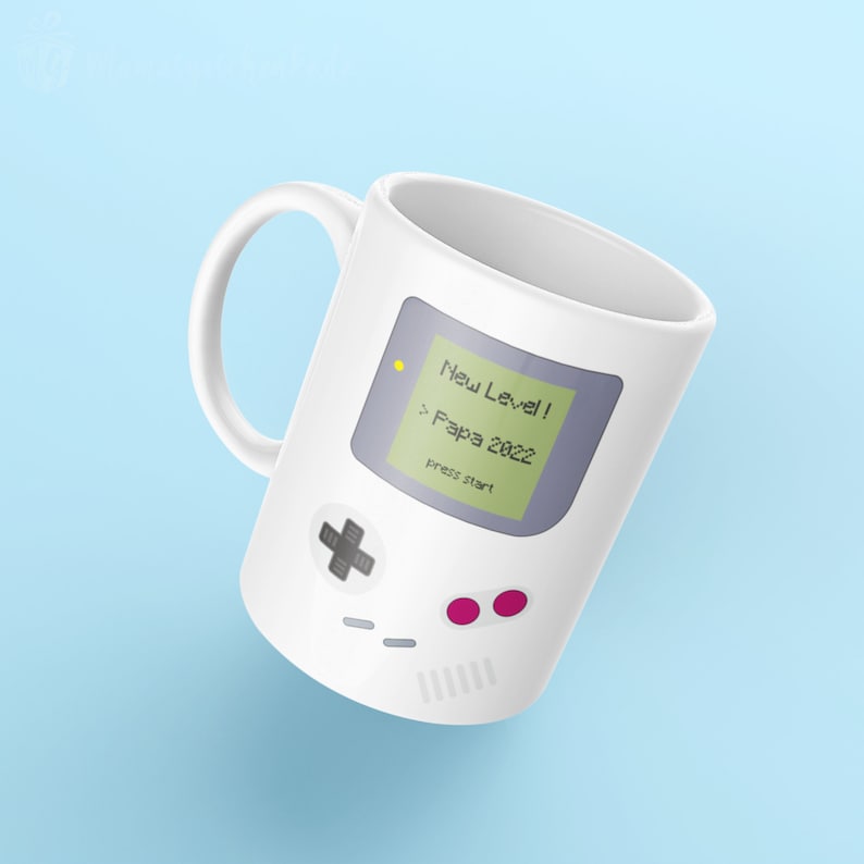 Puede incluir: Taza de cer&aacute;mica blanca con un gr&aacute;fico de una consola de videojuegos retro. La pantalla muestra "New Level! Papa 2022 press start".