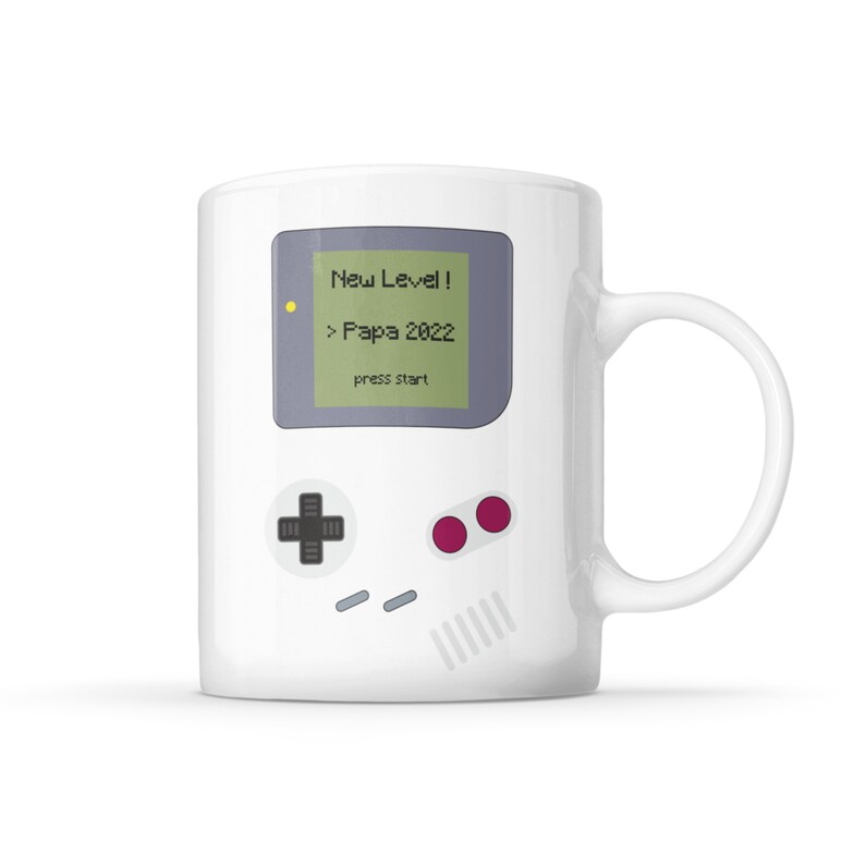 Puede incluir: Taza de cer&aacute;mica blanca con un gr&aacute;fico de una consola de videojuegos retro. La pantalla muestra "New Level! > Papa 2022 press start".