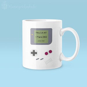 Puede incluir: Taza de cer&aacute;mica blanca con una ilustraci&oacute;n gris y verde de una consola de videojuegos retro con el texto "New Level! > Papa 2022 press start".