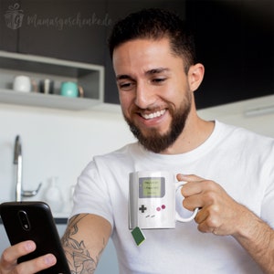 Puede incluir: Una taza de cer&aacute;mica blanca con un dise&ntilde;o negro y verde de una consola de videojuegos retro. La taza tiene un asa y el texto "Level 1" y "Press Start" en la parte delantera.