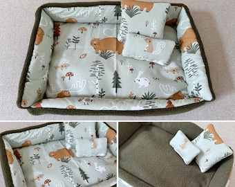 Puede incluir: Una cama para mascotas con temática de bosque. La cama tiene un interior verde claro con un estampado de osos, conejos y árboles. Incluye dos almohadas pequeñas y una almohada más grande, todas con diseños a juego. El borde de la cama es verde oscuro.