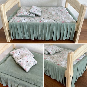 May include: A wooden doll bed with a green fleece blanket and a white floral patterned mattress and pillows. The bed has a fringe trim around the mattress.