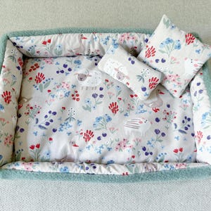 Peut inclure: Un lit rectangulaire pour animaux de compagnie avec un oreiller et un coussin assortis. Le lit a une bordure en polaire bleu clair et un intérieur à motifs floraux avec des moutons, des fleurs et des oiseaux. Les coussins ont le même motif.