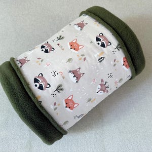 Peut inclure: Un lit cylindrique pour animaux de compagnie avec un imprimé d'animaux de la forêt. Le lit présente un tissu beige clair orné de ratons laveurs, de renards et de cerfs. Les bords sont bordés d'une polaire vert foncé. Le lit est conçu pour les petits animaux.