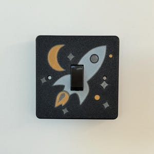 May include: Black light switch cover featuring a rocket ship, moon, and stars design. The rocket is grey, the moon and flames are gold, and the stars are white. The switch is black.