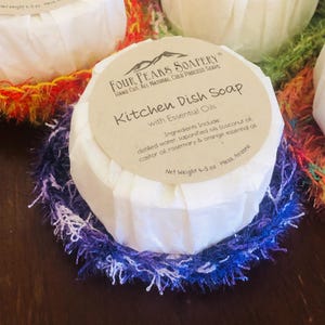 May include: Round, white wrapped kitchen dish soap with a label that reads "Four Peaks Soapery Kitchen Dish Soap with Essential Oils". The soap sits on a blue and purple crocheted scrubby. The net weight is 4-5 oz.