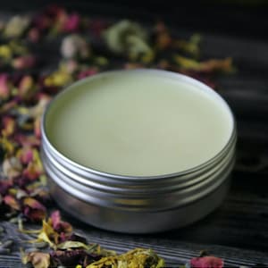 Könnte beinhalten: Eine kleine silberne Dose gefüllt mit einem hellgelben Balsam. Die Dose steht auf einer dunklen Holzoberfläche, umgeben von getrockneten Blütenblättern.