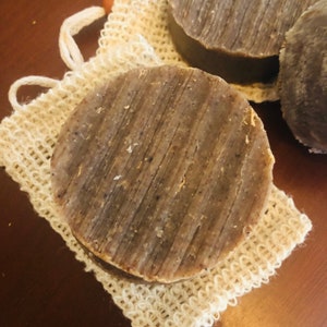 May include: Three round bars of brown soap with a textured surface, sitting on a white mesh bag.