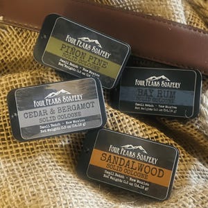 Könnte beinhalten: Vier kleine, rechteckige Dosen mit festem Kölnisch Wasser mit abgerundeten Ecken. Die Dosen sind schwarz mit weißem Text und einem Berglogo. Düfte sind Piñon Pine, Cedar & Bergamot, Bay Rum und Sandalwood. Jede Dose wiegt 14,18 g.