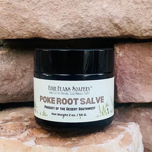 Könnte beinhalten: Ein schwarzes Glas Poke Root Salbe von Four Peaks Soapery, mit einem weißen Etikett. Auf dem Etikett steht "Product of the Desert Southwest" und "Netto-Gewicht 56 g.". Das Glas steht vor einem Hintergrund aus rotbraunem Stein.