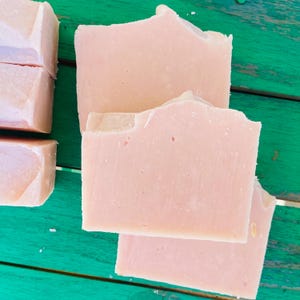 May include: Handmade soap bars in a pale pink colour are displayed on a green wooden surface. The rectangular bars have irregular edges, indicating a handcrafted origin. The soap's colour is uniform, and the texture appears smooth. The image suggests a natural, artisanal product.