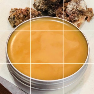 May include: A small, round, silver tin container filled with a thick, golden-brown balm or salve.